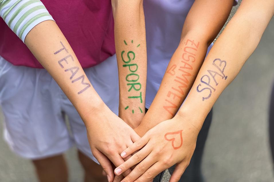 Vier Arme liegen übereinander, mit farbiger Schrift: „TEAM“, „SPORT“, „ZUSAMMEN“, „SPASS“ auf der Haut.